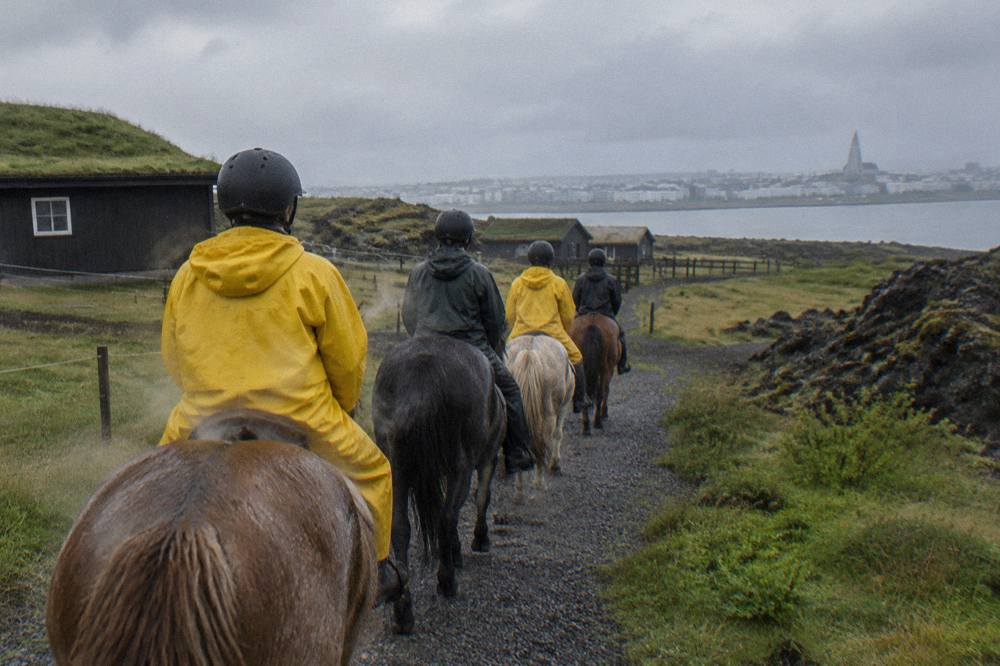 Beginner riders in helmets and rain gear walking single file on Icelandic horses along a gravel trail