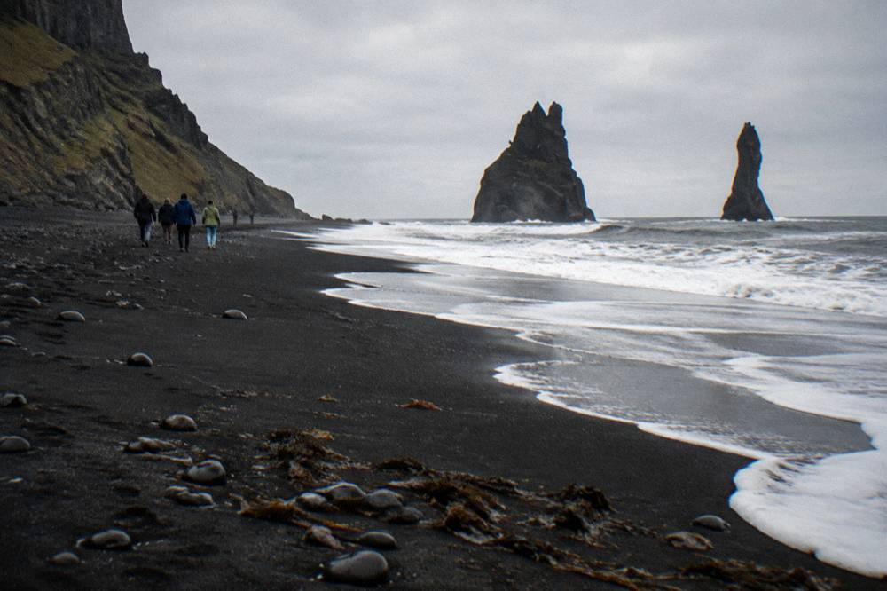 A wide black sand beach stretching toward tall basalt sea stacks under a grey sky