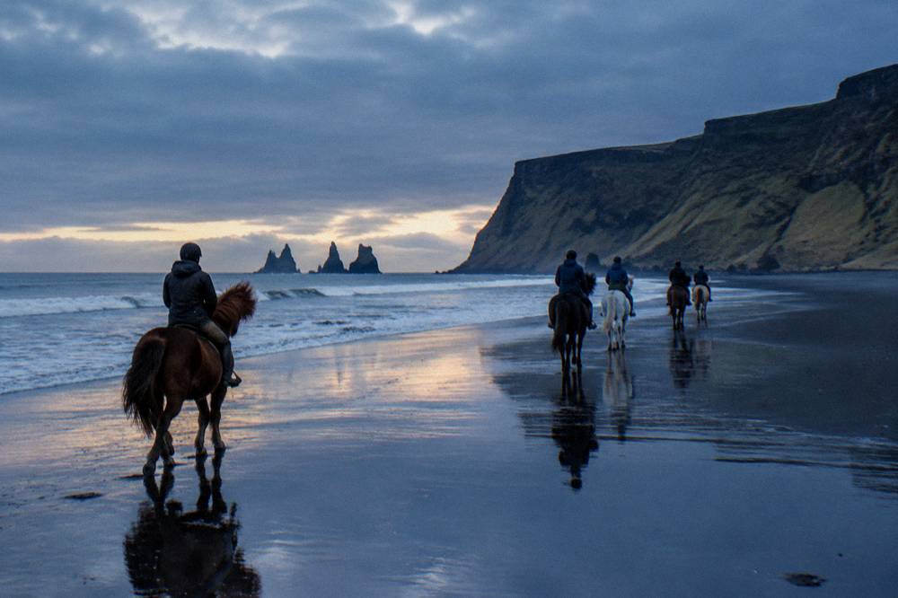 Riders on black sand under a wide evening sky with low light over the ocean