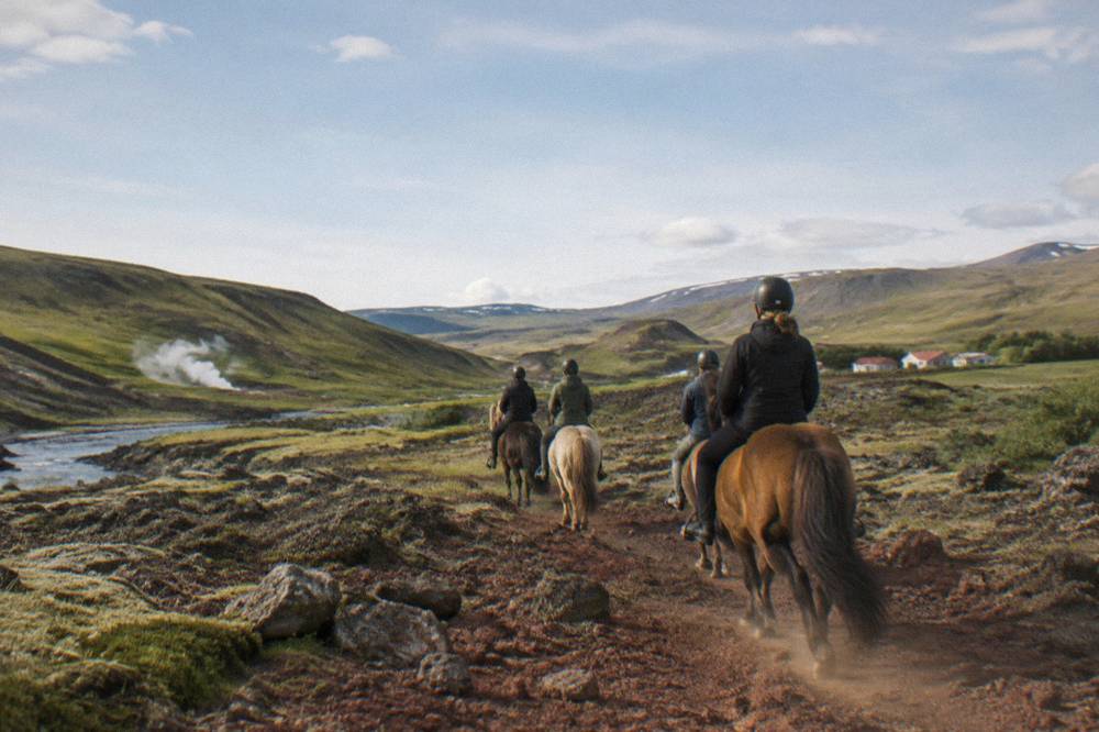 Riders on Icelandic horses crossing a red lava rock trail with moss patches under a wide sky