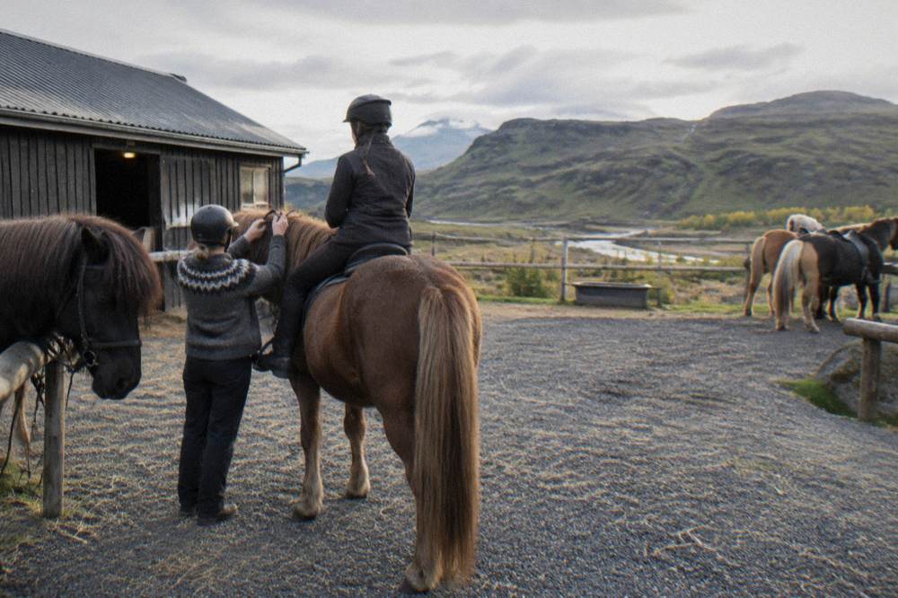A guide helping a mounted rider adjust the reins beside a stable yard with saddled Icelandic horses