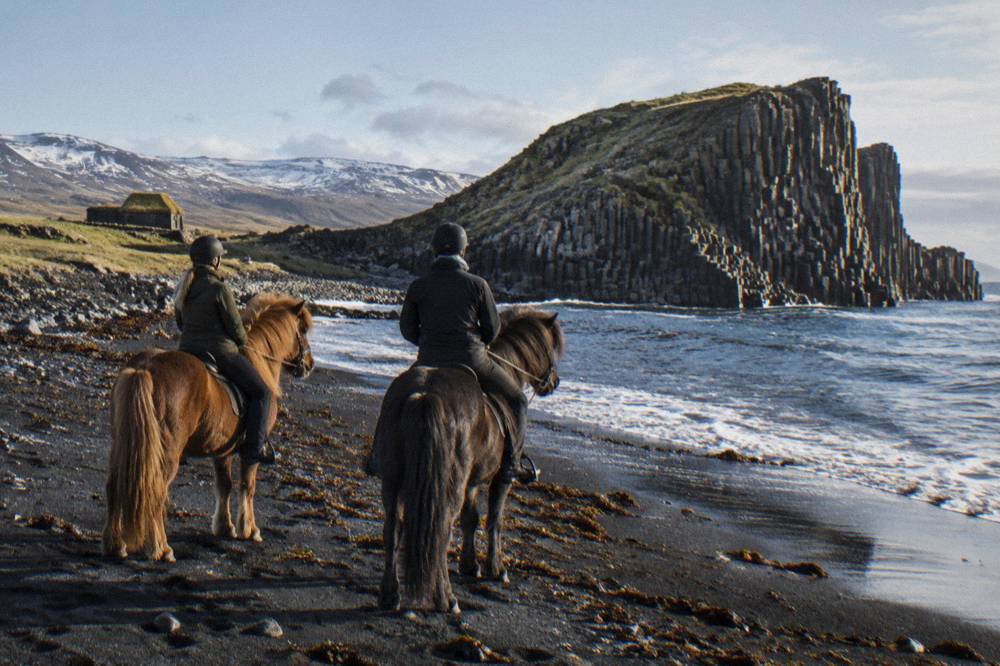 Two riders on Icelandic horses at the edge of a black sand beach with basalt columns in the distance
