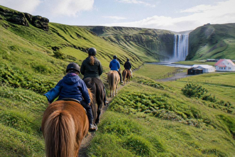 A small group of riders walking single file through green hills toward a distant waterfall