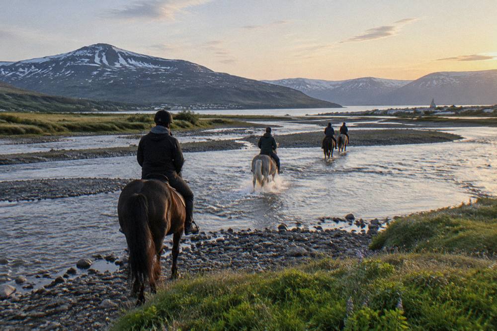 Riders crossing a wide river valley in northern Iceland under bright late evening sky