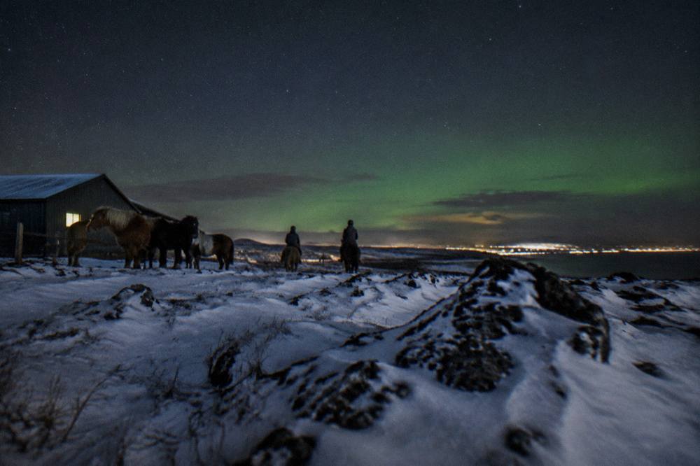 Snow covered lava field under a starry night sky with a faint green glow on the horizon